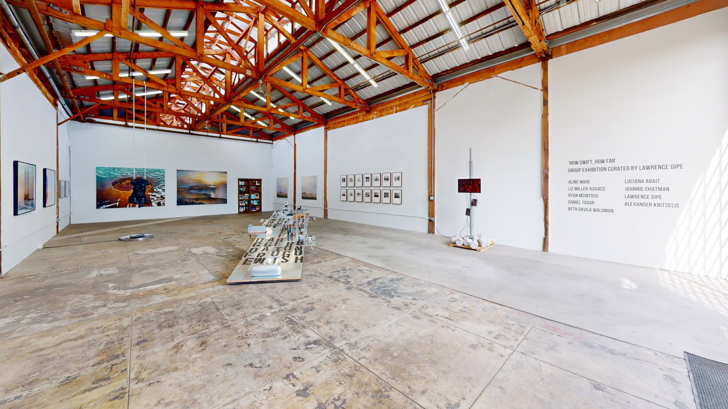 Wide gallery layout with floor-based installation pieces, wall art, and exhibition text under red-brown trusses.