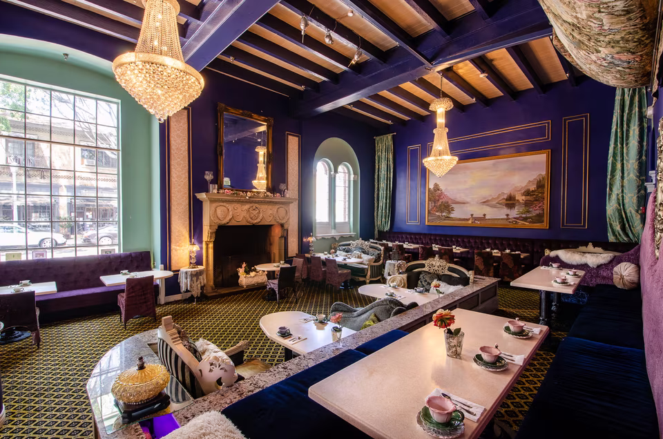 Deep-blue dining room with crystal chandeliers, carved fireplace, layered café seating, and exposed ceiling beams.