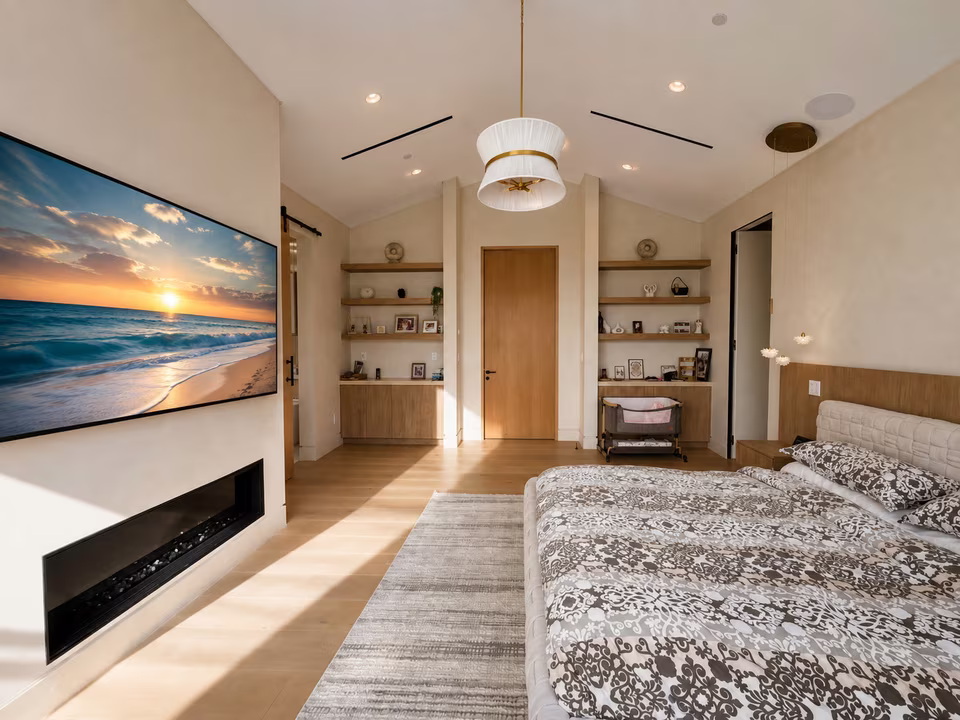 Primary bedroom sitting area with an upholstered bed, pale lounge chairs, warm wood flooring, and glass doors to a balcony.