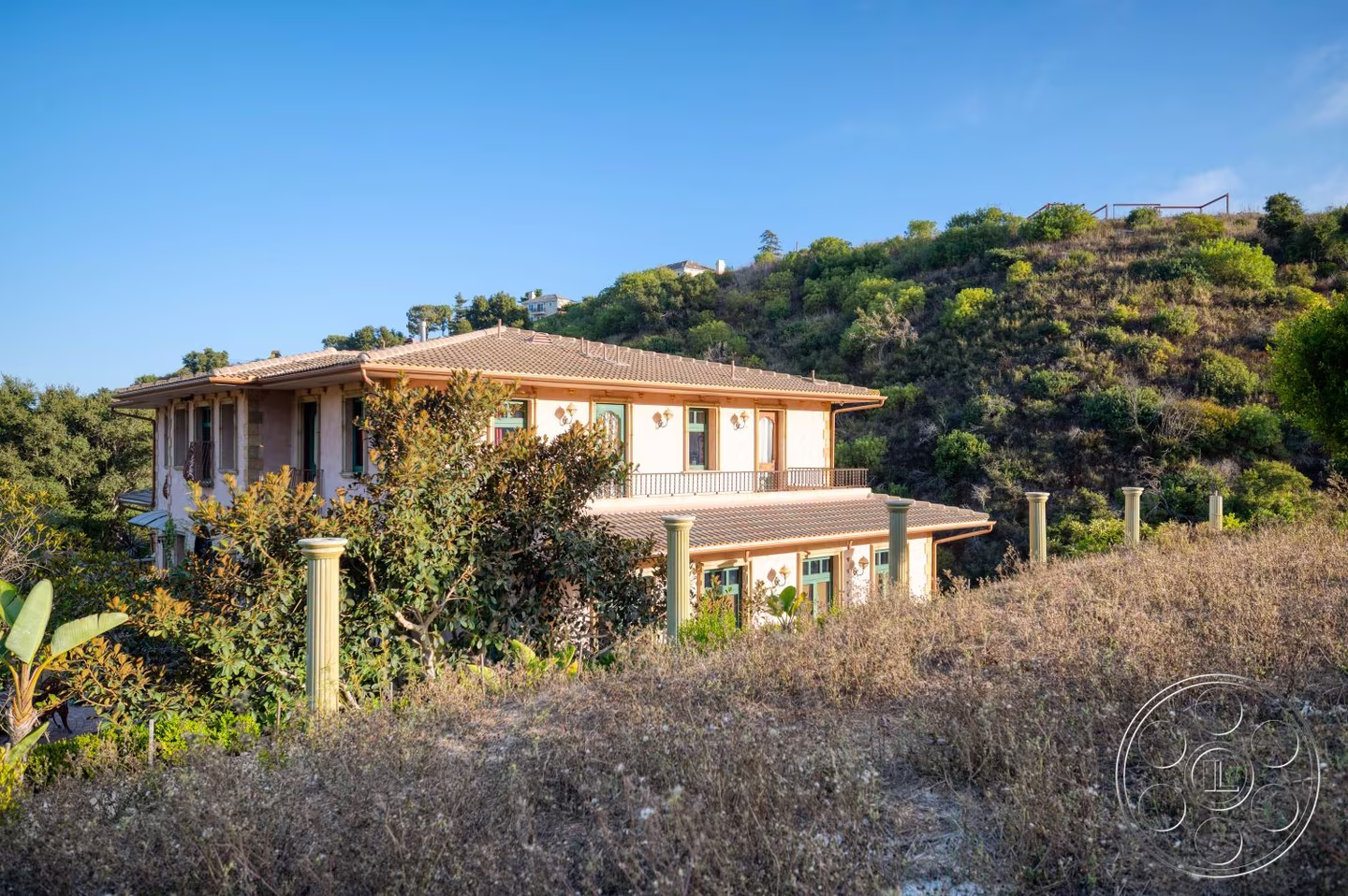 Wide exterior view of the villa roof, upper balcony, garden columns, and surrounding canyon hillside.